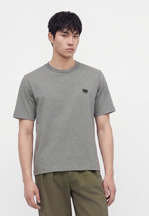 Jeune homme aux cheveux noirs courts portant un t-shirt gris uni à manches courtes et un pantalon vert olive sur un fond uni.