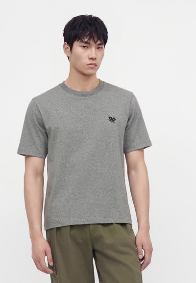 Jeune homme aux cheveux noirs courts portant un t-shirt gris uni à manches courtes et un pantalon vert olive sur un fond uni.