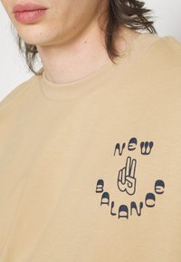 T-shirt en coton beige avec un logo noir rond portant le texte "NEW BALANCE" et un geste de la main au centre. Design à manches courtes.