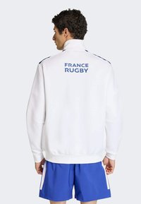 Biała kurtka na zamek z wysokim kołnierzem, niebieskie akcenty i napis "FRANCE RUGBY" na plecach. Dobranie do niebieskich szortów, gładka tekstura.