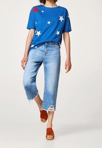 Camiseta azul con estrellas blancas y una estrella roja, combinada con unos vaqueros desgastados de color azul claro y sandalias de plataforma naranjas.