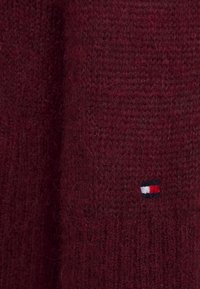 Bordeaux gebreide trui met een gestructureerde stof, voorzien van een klein geborduurd logo in rood, wit en blauw aan de onderzijde.
