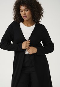 Cardigan lungo nero realizzato in tessuto morbido, con scollatura a V, apertura frontale e maniche lunghe. Indossato sopra una maglietta bianca e pantaloni neri.