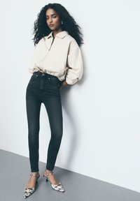 Beige cropped zip-up jack, high-waisted zwarte skinny jeans en bloemenprint pointed-toe hakken. Zachte texturen en een taillering fit.