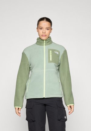 Fleece-takki vaaleanvihreänä, jossa on korkea kaulus, vetoketjullinen etuosa ja rintatasku. Ominaisuuksiin kuuluvat keltaiset vetoketjukoristeet ja pehmeä pinta.