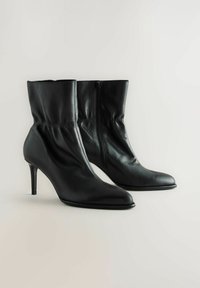 Mustad muste nahast keskmise pikkusega saapad teravate varvastega, kitsaste kontsadega ja külgzipperitega, paigutatud neutraalse tausta peale.