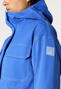 Μπλε κουκούλα από ανθεκτικό ύφασμα, με λευκές ραφές, τσέπη πατσαβούρα στο στήθος και λογότυπο στην μανσέτα.