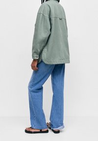 Person iført en lys grøn oversized jakke, løse blå jeans med frayed kanter og sorte flade sandaler, står imod en ensfarvet baggrund.
