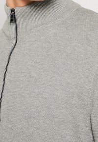 Närbild av en person som bär en ljusgrå stickad tröja med en svart dragkedja och en ribbad hög krage.