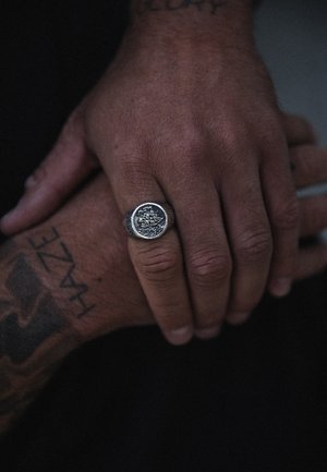 Silver signet ring with a detailed engraved design, featuring a textured band. Two hands are clasped, one tattooed.