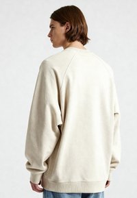 Jeune personne aux cheveux bruns mi-longs, tournée vers la gauche, portant un sweat-shirt crème surdimensionné et un jean bleu sur un fond uni.