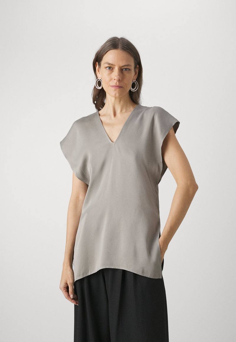 Light gray v-neck top with short sleeves, made from a smooth fabric. Features a relaxed fit and rounded hem, paired with black trousers.