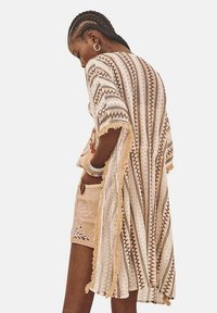 Kimono hasta la rodilla en un suave beige con intrincados patrones geométricos coloridos, bordes con flecos y mangas cortas; hecho de una tela ligera.