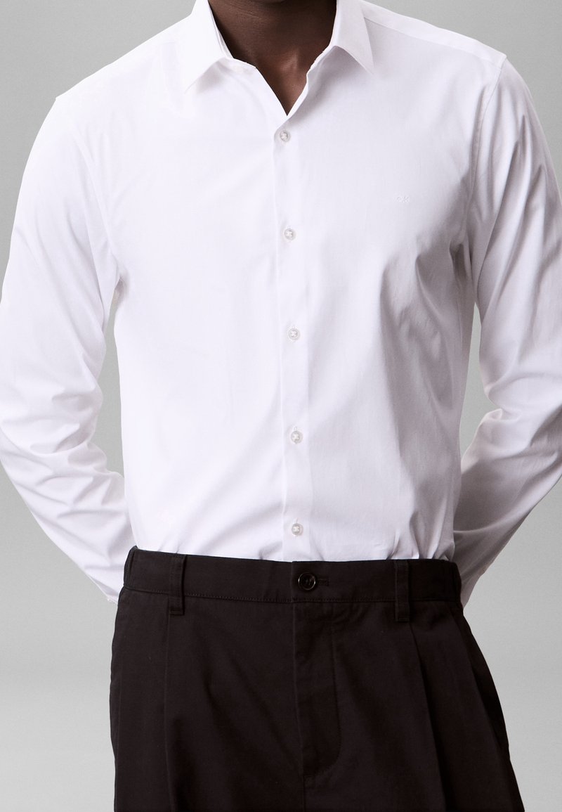 Homme portant une chemise blanche cintrée à manches longues boutonnée, rentrée dans un pantalon noir taille haute, sur un fond uni.