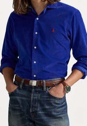 Mężczyzna ubrany w niebieską koszulę na guziki z sztruksu z podwiniętymi rękawami, brązowy skórzany pasek, zegarek na nadgarstku i niebieskie dżinsy, z rękami w kieszeniach.