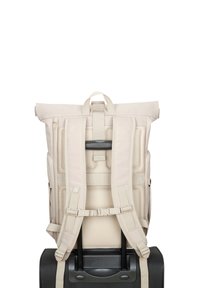 Sac à dos beige à rabat sécurisé sur la poignée d'une valise à roulettes noire, présentant des détails sur les bretelles rembourrées et le panneau arrière.