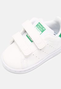 Bílé tenisky Adidas Stan Smith se zeleným logem na jazyku a dvěma suchými zipy na světlém pozadí.