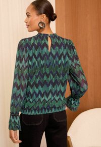Blusa a righe chevron verde e blu con maniche lunghe, texture plissettata, schiena a chiave e polsini arricciati con dettagli in perle.