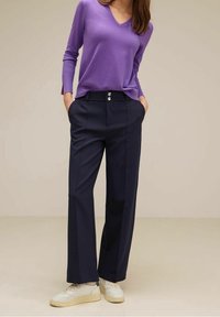 Femme portant un pull col en V lavande, un pantalon large taille haute bleu marine avec double boutons, et des baskets blanches, debout les mains dans les poches.