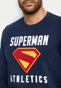 Granatowy sweatshirt z dużym wyhaftowanym logo Supermana w czerwonym i żółtym kolorze. Na tekście widnieje napis "SUPERMAN" oraz "ATHLETICS" w białym kolorze. Wykonany z miękkiej bawełny.