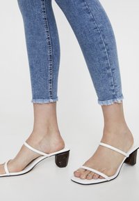 Sandales blanches à talons avec deux fines brides sur le pied et un talon en bois marron épais. Associées à un pantalon en denim bleu avec un ourlet effiloché.