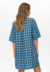 Donna con capelli lunghi che indossa un vestito a quadri blu e nero, a maniche corte e largo, in piedi con la schiena rivolta verso la macchina fotografica.