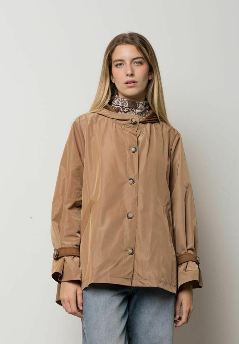 Mujer con impermeable marrón claro abotonado sobre una camiseta de cuello alto con estampado y jeans azul claro, de pie frente a un fondo liso.