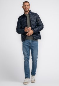 Marineblau gesteppte Steppjacke mit Stehkragen, Reißverschluss vorne und gerippten Bündchen, kombiniert mit einem grauen strukturierten Pullover und hellblauen Jeans.