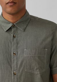 Korte mouwen, olijfgroene shirt gemaakt van gestructureerde stof. Heeft een button-down kraag, knopen aan de voorkant en een enkele borstzak met witte stiksels.