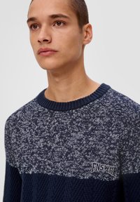 Marineblauer und grauer Strickpullover mit strukturiertem Muster und geripptem Kragen. Mit einem Logo-Patch auf der linken Seite.