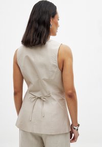 Beige ärmlös topp med en figurnära siluett, bakknutning och slät textur. Den släta tyget och det enkla designen skapar en minimalistisk look.