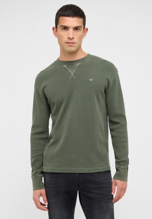 Zaļa adīta krekla ar garām piedurknēm, teksturētu rakstu, apaļu apkakli un nelielu logotipa uzšuvumu krūtīs. Kombinēts ar tumši pelēkām biksēm.