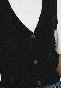 Schwarze Strickweste mit tiefem V-Ausschnitt, strukturiertem Stoff und vier Knopfverschlüssen vorne. Trägt man über einem weißen Hemd.