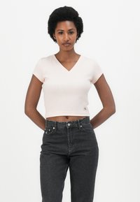 Crop top a maniche corte di colore rosa chiaro con scollo a V e texture a coste, abbinato a jeans in denim scuro a vita alta. Piccolo logo sull'orlo.