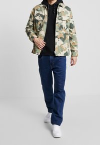 Veste à motif camouflage en vert et beige portée sur un sweat à capuche noir, associée à un jean bleu et des baskets blanches, avec deux poches poitrine visibles.