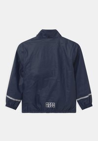 Navy waterproof jacket with a high collar, elastic cuffs, and reflective stripes. Features a textured panel with circular cutouts at the back.