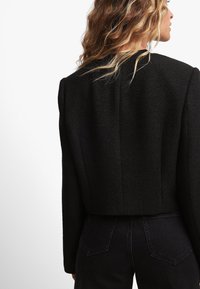 Mustreeritud kangast must lühike jakk, millel on rõhutatud õlgade ja puhas joonis. Seljal on tagasihoidlikud õmblused ja struktureeritud siluett.