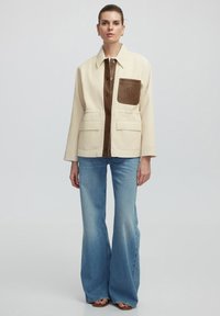 Beige gewatteerde jas met een bruine leren voorpaneel en zak, met twee voetzakken. Gecombineerd met uitlopende blauwe jeans.