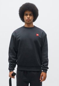 Sudadera de algodón negra con puños y dobladillo acanalados, que presenta un pequeño logo rojo en el pecho. El modelo sostiene una correa, de pie contra un fondo blanco.