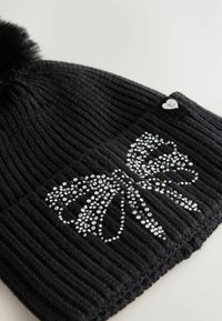 Black knitted beanie with a ribbed texture, featuring a silver rhinestone bow design on the front and a faux fur pom-pom on top.