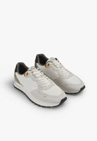 Zapatillas blancas y grises que combinan materiales de malla y ante, con suelas de goma negras y acentos en naranja y marrón en el talón.