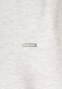 Tejido de color gris claro con una pequeña etiqueta metálica plateada con "SKILODX" grabado, situada en la parte inferior derecha.