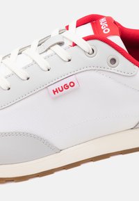 Graue und weiße Sneaker mit einem strukturierten Canvas-Obermaterial, roten Akzenten, Gummisohle und einem auffälligen "HUGO"-Logoschild an der Seite.