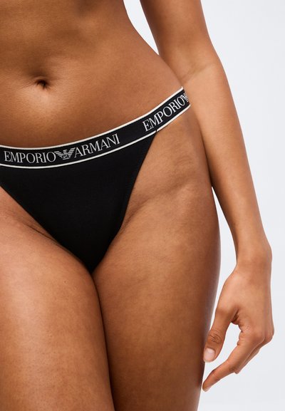 Gros plan d'une personne portant des sous-vêtements noirs Emporio Armani avec une ceinture blanche à logo, montrant la taille et le haut des cuisses.