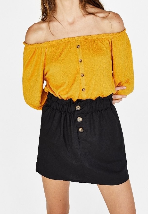 Femme portant un haut jaune moutarde à épaules dénudées avec des boutons et une jupe noire taille haute avec des détails boutonnés.