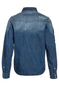 Camisa de mezclilla en azul medio con un acabado ligeramente desgastado. Cuenta con un cuello abotonado, mangas largas y parches en los codos en ambos brazos.