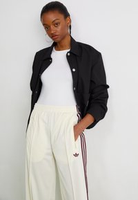 Schwarze Knopfleistenjacke über einem weißen Hemd, kombiniert mit beigen Hosen mit burgunderfarbenen Streifen und einem Adidas-Logo. Einfaches, modernes Design.
