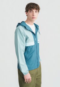 Νεαρός άνδρας με κατσαρά μαλλιά που φοράει ζακέτα με κουκούλα σε τιρκουάζ και ανοιχτό γαλάζιο με φερμουάρ και ελιές πράσινο παντελόνι, στέκεται μπροστά σε απλό λευκό φόντο.