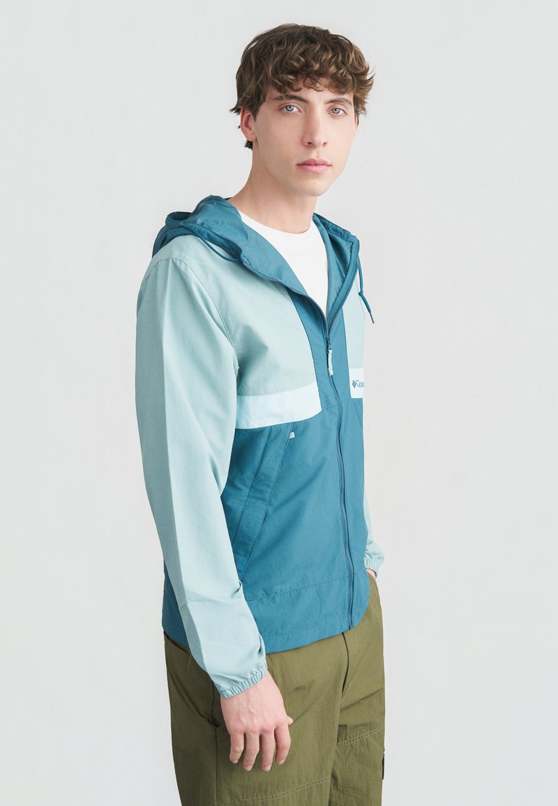 Νεαρός άνδρας με κατσαρά μαλλιά που φοράει ζακέτα με κουκούλα σε τιρκουάζ και ανοιχτό γαλάζιο με φερμουάρ και ελιές πράσινο παντελόνι, στέκεται μπροστά σε απλό λευκό φόντο.