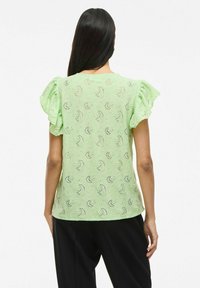 Top verde in pizzo con maniche a volant, caratterizzato da motivi delicati e una finitura testurizzata, abbinato a pantaloni neri; vista posteriore mostrata.